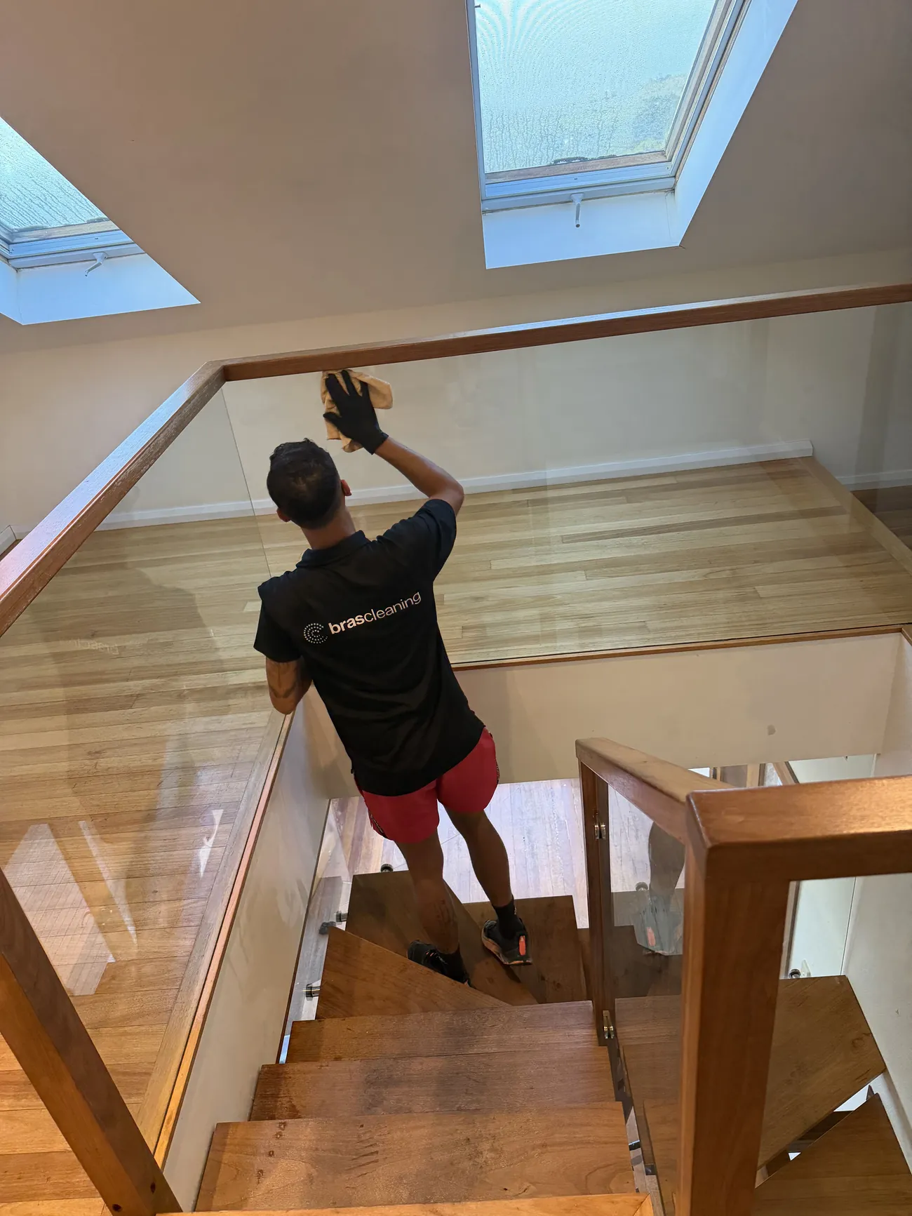 Cleaner wiping a glass balustrade on a timber staircase under a skylight